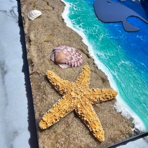 Puede incluir: Una pieza de arte de resina que representa una escena de playa con una estrella de mar, conchas marinas y una silueta de ballena en el oc&eacute;ano. El oc&eacute;ano es azul y verde con olas de espuma blanca. La arena es marr&oacute;n y texturizada.