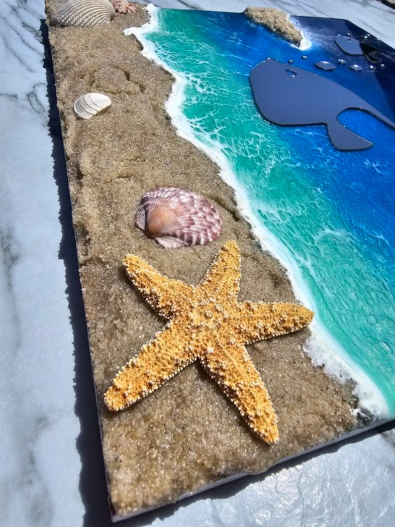 Puede incluir: Una pieza de arte de resina que representa una escena de playa con una estrella de mar, conchas marinas y una silueta de ballena en el oc&eacute;ano. El oc&eacute;ano es azul y verde con olas de espuma blanca. La arena es marr&oacute;n y texturizada.