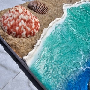 May include: A coastal-themed resin art piece featuring a beach scene. The artwork includes a sandy area with seashells and a turquoise and blue ocean with foamy white waves. The large seashell is red and white.