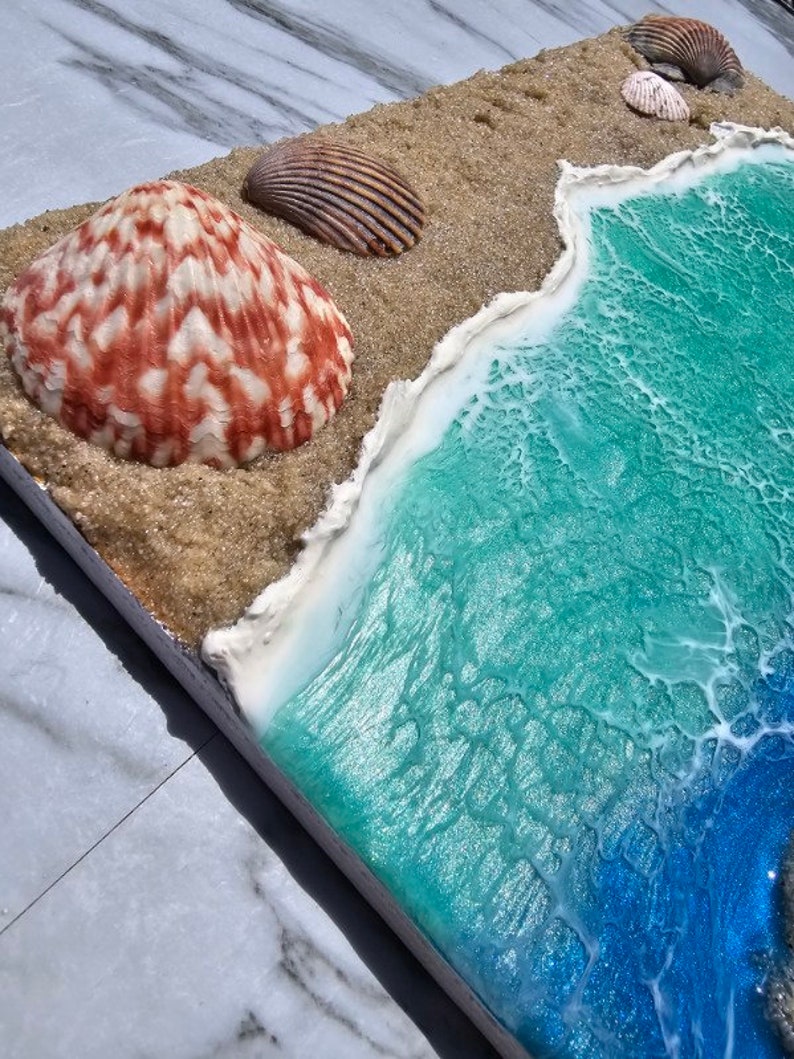 May include: A coastal-themed resin art piece featuring a beach scene. The artwork includes a sandy area with seashells and a turquoise and blue ocean with foamy white waves. The large seashell is red and white.