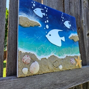 Puede incluir: Una pieza de arte de resina azul y verde con siluetas de peces blancas y textura de arena. La pieza presenta conchas marinas y una estrella de mar.