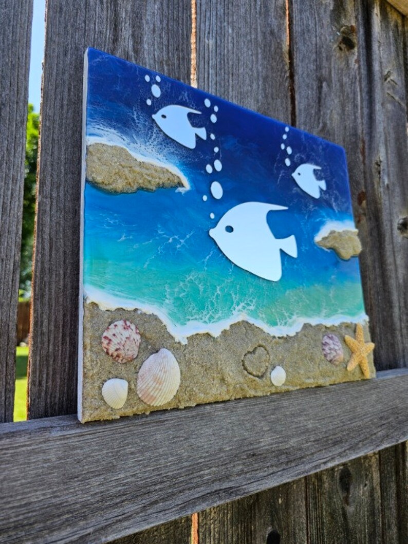Puede incluir: Una pieza de arte de resina azul y verde con siluetas de peces blancas y textura de arena. La pieza presenta conchas marinas y una estrella de mar.