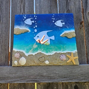 Puede incluir: Una pieza de arte de resina azul y blanca con una escena de playa. La pieza presenta tres peces blancos, un coraz&oacute;n y conchas en una playa de arena.