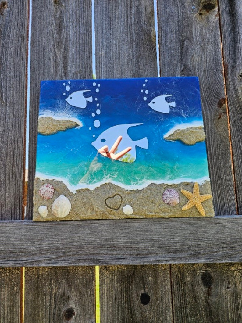 Puede incluir: Una pieza de arte de resina azul y blanca con una escena de playa. La pieza presenta tres peces blancos, un coraz&oacute;n y conchas en una playa de arena.