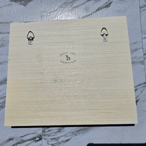 Puede incluir: Una obra de arte enmarcada en madera clara con dos ganchos de metal para colgar en la parte posterior. La obra de arte tiene un logotipo en blanco y negro que dice "Cerie Art Emporium" con una B estilizada.