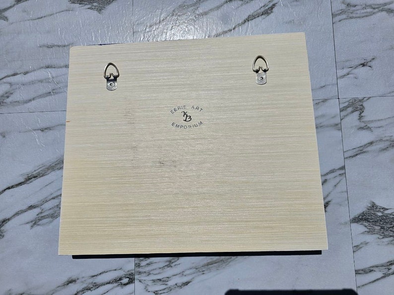 Puede incluir: Una obra de arte enmarcada en madera clara con dos ganchos de metal para colgar en la parte posterior. La obra de arte tiene un logotipo en blanco y negro que dice "Cerie Art Emporium" con una B estilizada.