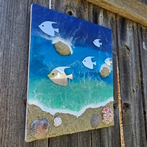 May include: A rectangular wall art piece depicting an ocean scene. The artwork features a blue and turquoise ocean with white fish, seashells, and a sandy beach. The art is mounted on a white frame and is displayed against a wooden fence.
