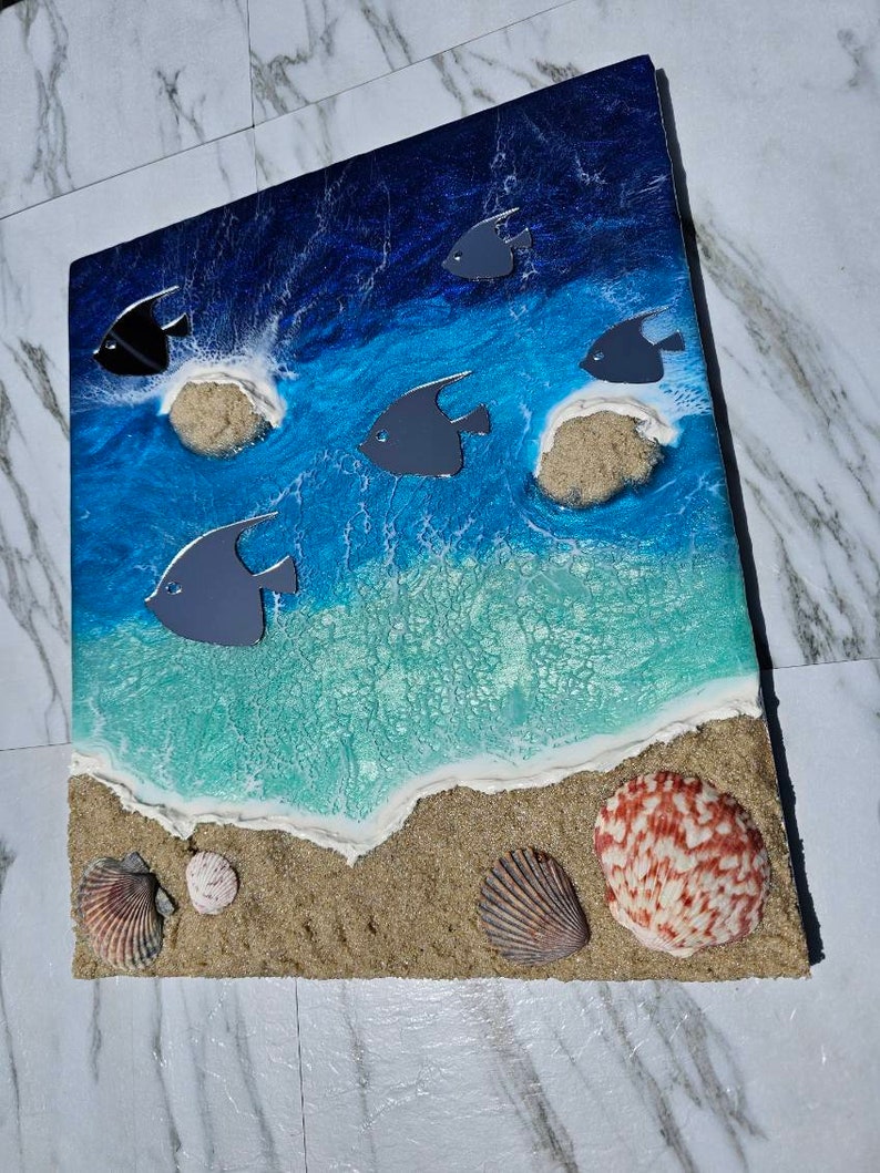 May include: Ocean-themed artwork featuring a beach scene with blue and turquoise water, sand, and seashells. Gray fish swim above the waves. The artwork includes a textured sand and shell border.