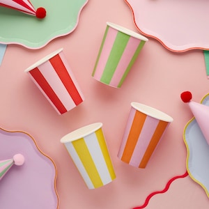 Puede incluir: Vista aérea de suministros para fiestas coloridos sobre un fondo rosa. Incluye vasos de papel a rayas en rojo, verde, naranja y amarillo, junto con platos y sombreros de fiesta a juego. Un arreglo festivo y alegre.