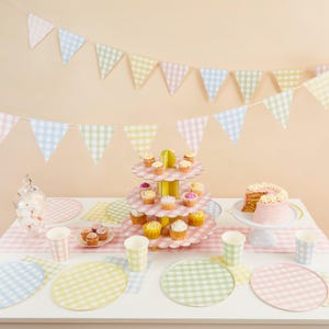 Può includere: Un'apparecchiatura da tavola festosa con decorazioni a quadretti pastello. Include un portacupcake a più livelli, una torta su un piedistallo e una ghirlanda. Lo schema dei colori presenta rosa, giallo, verde e blu.