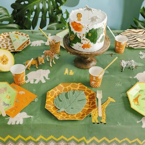 May include: A festive safari-themed party table setting with a decorated cake, animal figurines, and patterned plates and cups. The green tablecloth features a repeating geometric pattern and animal illustrations.