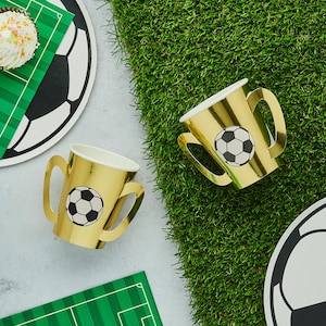 Puede incluir: Vasos de papel dorados con asas, cada uno con un diseño de balón de fútbol en blanco y negro. Los vasos están colocados sobre una superficie con césped verde artificial y artículos para fiestas temáticas de fútbol.