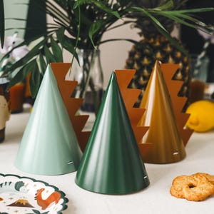 May include: Three party hats in green, brown and teal with a dinosaur design. The hats are on a white table with a plate and biscuits.