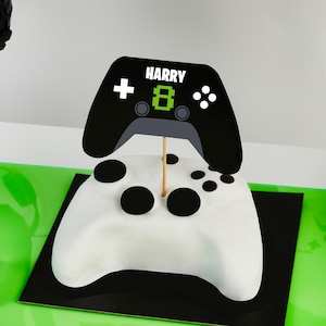 May include: A white cake shaped like a video game controller, topped with a black controller-shaped decoration. The decoration has the name "HARRY" and the number "8" on it. The cake sits on a black base.