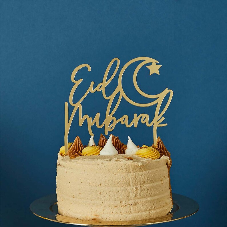May include: A gold cake topper with the words "Eid Mubarak" and a crescent moon and star design. The topper is on top of a frosted cake with brown and yellow frosting.