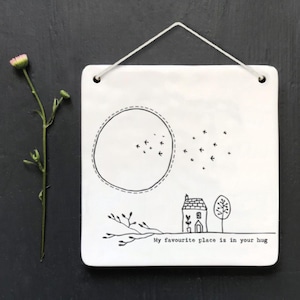 Può includere: Arte da parete quadrata in ceramica bianca con un disegno a tratto nero di una casa, un albero e uccelli. Il testo "My favourite place is in your hug" è in basso. A sinistra c'è un fiore rosa e verde.