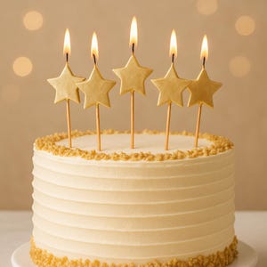 Peut inclure: Un gâteau décoré de cinq bougies dorées en forme d'étoile. Le gâteau est recouvert d'un glaçage blanc avec des rainures horizontales et une bordure de paillettes dorées. Les bougies sont allumées avec une flamme jaune.