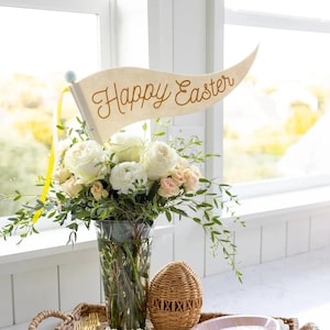 May include: A vase of white and peach roses with green foliage, and a banner reading "Happy Easter" in gold script. The arrangement is in a clear glass vase. The display includes a woven basket and a decorative plate.