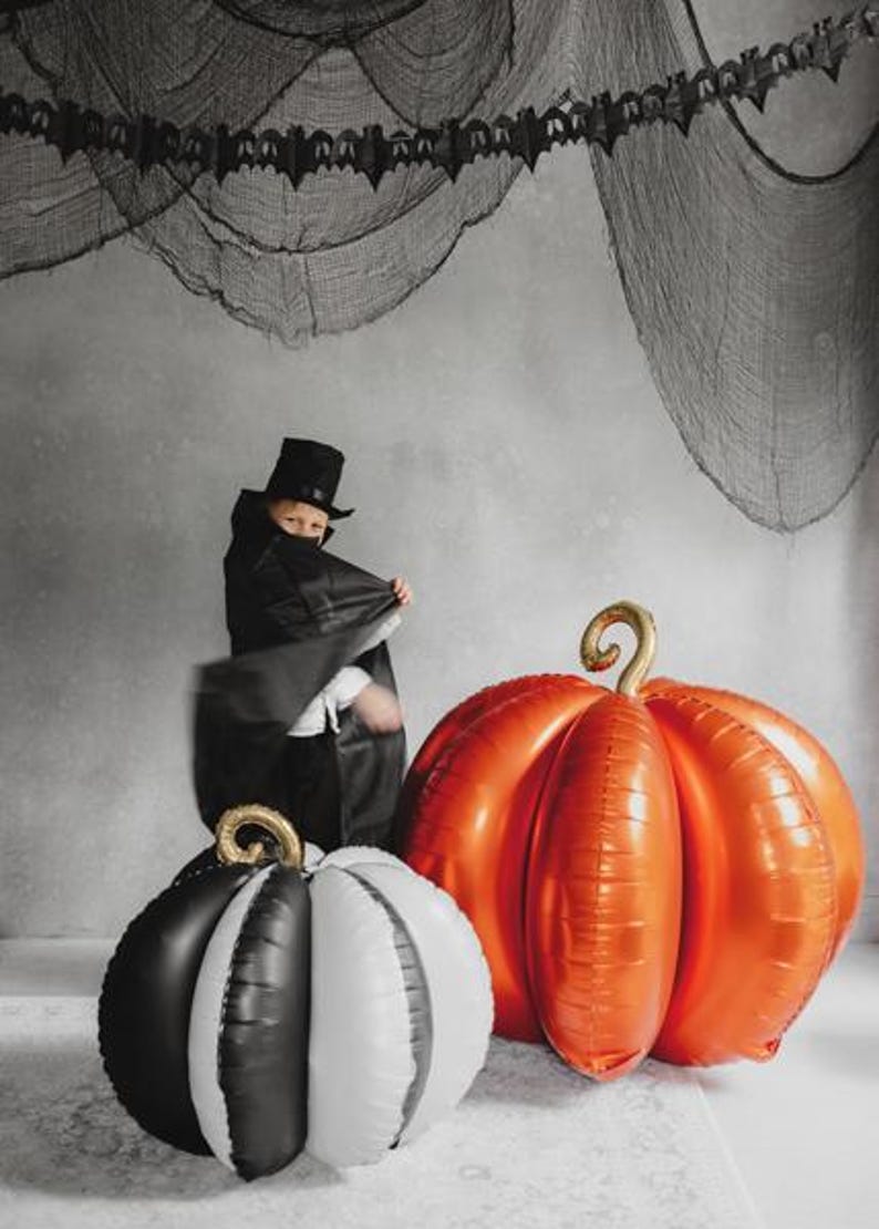 May include: A Halloween-themed image featuring a child in a black costume and top hat. Two large pumpkin-shaped balloons, one orange and one black and white, are in the foreground. A black bat garland hangs in the background.