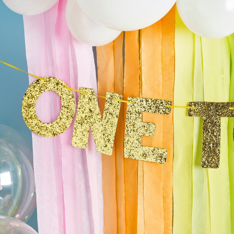 Pu&ograve; includere: Uno striscione di glitter dorati con la parola "ONE" in grandi lettere. Lo striscione &egrave; appeso su uno sfondo di nastri di carta velina rosa, arancio e giallo.