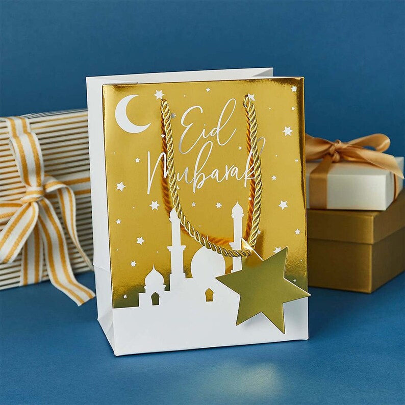 May include: A white gift bag with a gold foil design. The bag features a crescent moon, stars, and a mosque silhouette. The text "Eid Mubarak" is printed in white on the bag. A gold star is attached to the bag's handle.