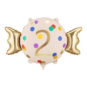 Op de afbeelding: Een lichtroze snoepvormige ballon met het nummer 2 in goud. De ballon is versierd met kleurrijke stippen en gouden folie accenten. De ballon is ontworpen voor een tweede verjaardag.