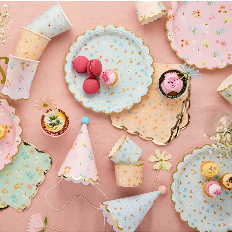 Puede incluir: Un conjunto de art&iacute;culos para fiestas con un dise&ntilde;o floral. Incluye platos, vasos, envoltorios para cupcakes y gorros de fiesta en colores pastel. Los platos tienen un borde festoneado y ribete dorado. Macarons y cupcakes est&aacute;n colocados en los platos.