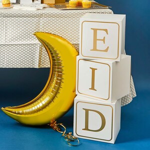 May include: White cardboard boxes stacked to spell "EID" with gold lettering. A gold crescent moon balloon is in the foreground.
