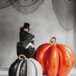 May include: A Halloween scene featuring three large pumpkin-shaped balloons. One is orange, another is black and white, and the third is black and white. A child in a black costume and top hat stands near the balloons. A black bat garland hangs in the background.