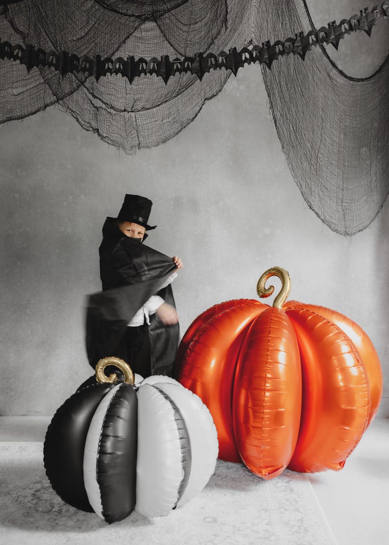 May include: A Halloween scene featuring three large pumpkin-shaped balloons. One is orange, another is black and white, and the third is black and white. A child in a black costume and top hat stands near the balloons. A black bat garland hangs in the background.