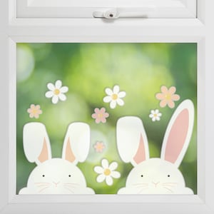 May include: Two white bunny rabbits with pink ears peeking through a window with a blurred green background.  The window is decorated with white and pink flowers.