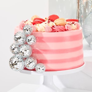 Peut inclure: Un g&acirc;teau &agrave; &eacute;tages rose et blanc d&eacute;cor&eacute; de macarons et surmont&eacute; d'ornements en forme de boule disco argent&eacute;e. Le g&acirc;teau est sur un support &agrave; g&acirc;teau blanc.