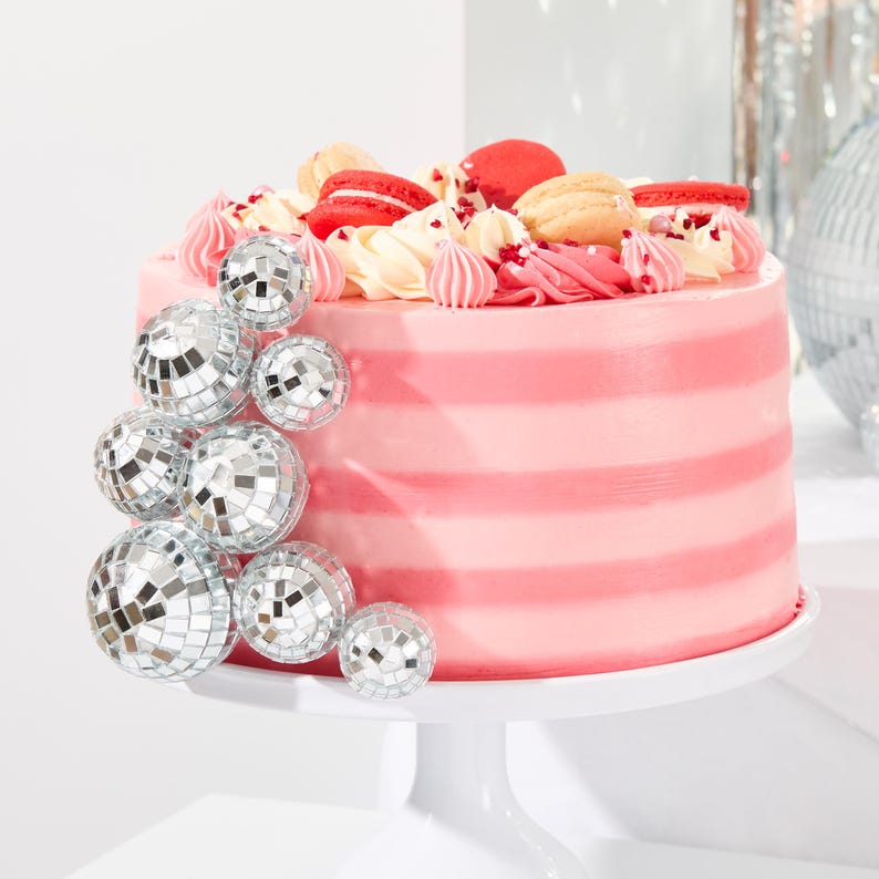Peut inclure: Un g&acirc;teau &agrave; &eacute;tages rose et blanc d&eacute;cor&eacute; de macarons et surmont&eacute; d'ornements en forme de boule disco argent&eacute;e. Le g&acirc;teau est sur un support &agrave; g&acirc;teau blanc.