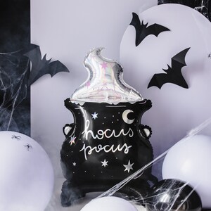 May include: A black Halloween cauldron-shaped balloon with the words "hocus pocus" in white script. A silver, flame-shaped balloon sits atop the cauldron. The background includes black bat cutouts, white balloons, and spiderwebs.