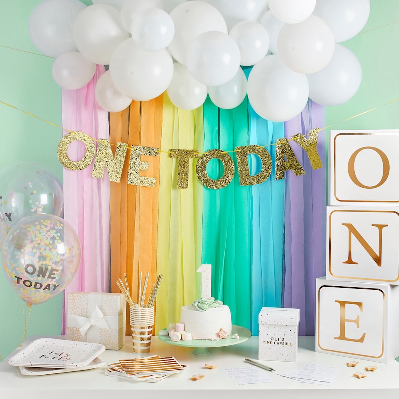 Pu&ograve; includere: Un set di decorazioni per una festa di primo compleanno con uno striscione di glitter dorato che dice "One Today", palloncini bianchi, festoni arcobaleno e blocchi dorati e bianchi che compongono la parola "ONE".