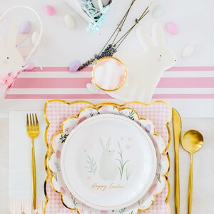 May include: Easter table setting with a white and gold scalloped plate, a pink gingham placemat, and gold cutlery. The plate features a bunny illustration and the text "Happy Easter". Decorative elements include a bunny-shaped plate and pastel-coloured sweets.