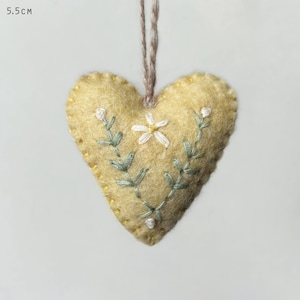 Puede incluir: Adorno en forma de corazón de fieltro amarillo, de 5,5 cm de largo. Presenta diseños florales bordados en blanco y verde. Se adjunta una cuerda marrón para colgarlo. El adorno tiene un borde cosido.