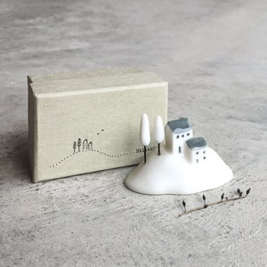 Op de afbeelding: Een klein wit keramiek beeldje van een heuvel met twee huizen en drie bomen. Het beeldje staat op een bruine doos met het woord "Hillside" erop.