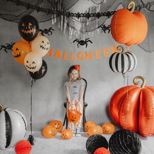 May include: A Halloween-themed scene with orange and black pumpkin balloons, spider decorations, and a banner that reads "HALLOWEEN." A young person is holding a pumpkin-shaped bucket. The scene is festive and colorful.