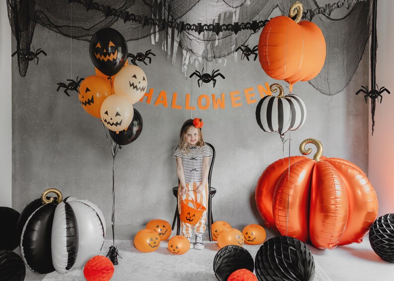 May include: A Halloween-themed scene with orange and black pumpkin balloons, spider decorations, and a banner that reads "HALLOWEEN." A young person is holding a pumpkin-shaped bucket. The scene is festive and colorful.