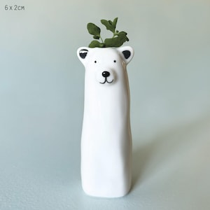 May include: A white ceramic polar bear vase, measuring 6 x 2 cm, with black detailing for the nose, eyes, and ears. The vase contains green foliage. The vase has a smooth, glossy finish and a playful design.