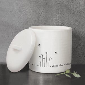 Puede incluir: Tarro de cerámica blanco con tapa, con un diseño minimalista de flores y abejas en línea negra. El texto "...Keep the change" está impreso en el tarro. El tarro está sobre una superficie oscura.