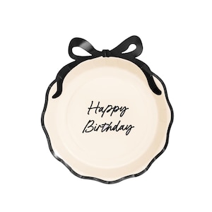 May include: A cream-coloured paper plate with a black scalloped edge and a black bow at the top. The words "Happy Birthday" are written in black script in the centre of the plate.
