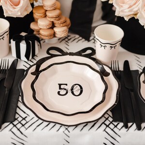 Può includere: Piatti di carta bianco e nero con un design a fiocco nero e il numero 50 con la parola "hello" scritta sotto. I piatti sono su una tovaglia a scacchi bianco e nero.