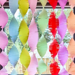 Könnte beinhalten: Eine farbenfrohe, rautenförmige Papiergirlande in Rosa, Blau, Gelb, Grün, Rot und Orange hängt vor einem Fenster. Die Girlande besteht aus geschichtetem, gefranstem Papier und erzeugt einen strukturierten, dekorativen Effekt. Das Fenster zeigt Bäume und einen Zaun.