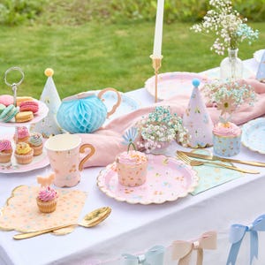 Puede incluir: Una mesa de fiesta decorada con platos estampados florales, cupcakes y una tetera azul de panal. La escena incluye sombreros de fiesta, macarons y cubiertos dorados, creando un ambiente caprichoso y alegre.