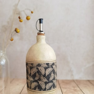 Op de afbeelding: Een keramische olijfolie fles met een zwart-wit verenpatroon en een bijpassend keramisch schaaltje. De fles heeft een kurkstop met een metalen giettuit.