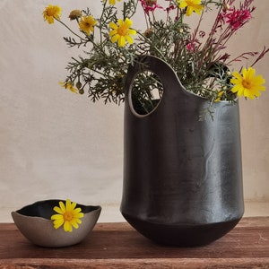 Könnte beinhalten: Eine schwarze Keramikvase mit einem einzigartigen Ausschnittdesign hält einen Strauß gelber und rosa Blumen. Eine kleine graue Schale mit einer gelben Blume steht auf einer Holzoberfläche.
