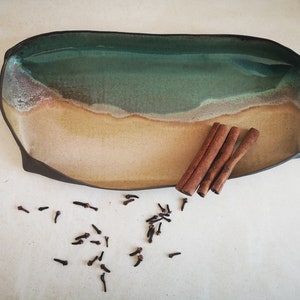 Puede incluir: Una bandeja de servir de cer&aacute;mica con un esmalte verde y marr&oacute;n, que recuerda a una escena de playa. La bandeja est&aacute; llena de palitos de canela y clavos.