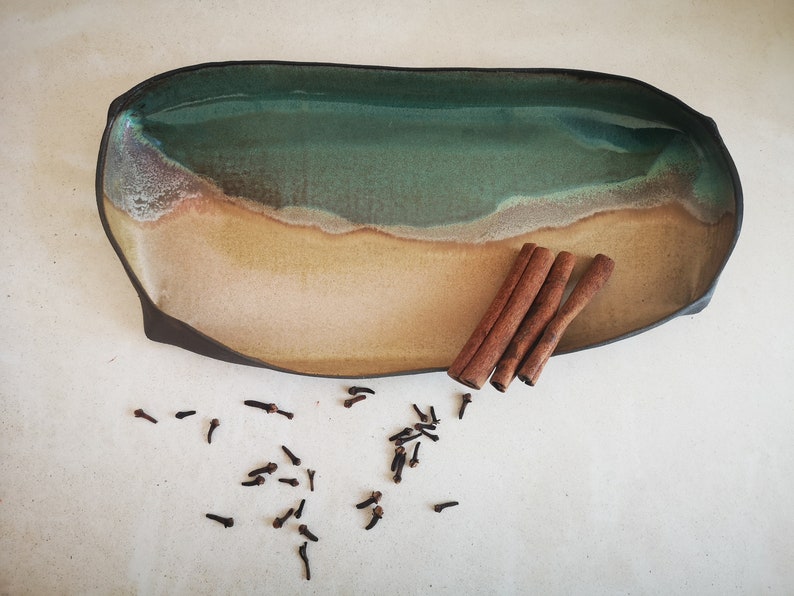 Puede incluir: Una bandeja de servir de cer&aacute;mica con un esmalte verde y marr&oacute;n, que recuerda a una escena de playa. La bandeja est&aacute; llena de palitos de canela y clavos.